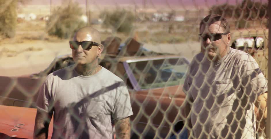 Two men stand near hot rods by a fence