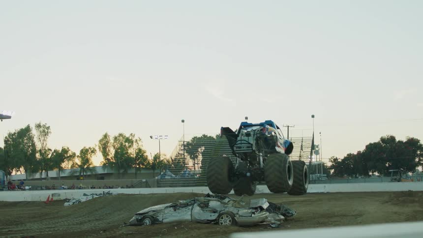 A monster truck rolls over a pile of wrecked cars in a stadium at dusk