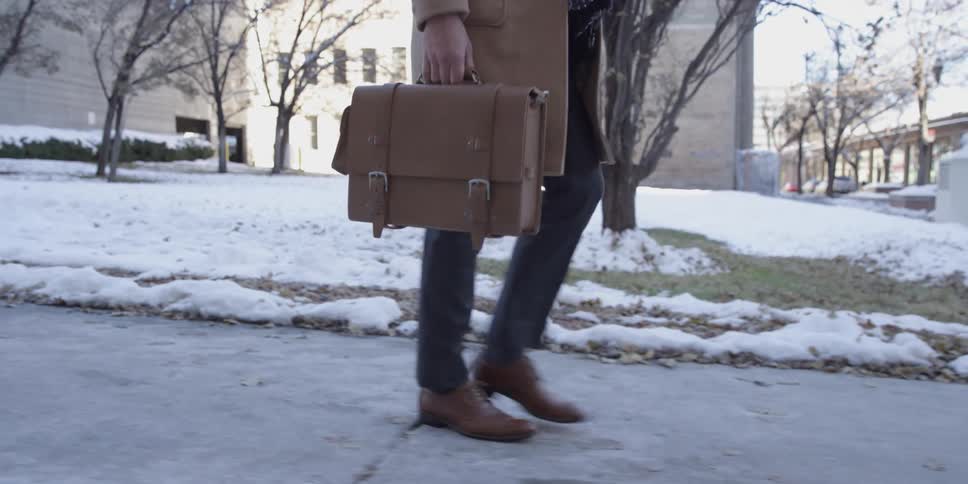 Someone is walking on the sidewalk next to snow covered grass holding a leather briefcase
