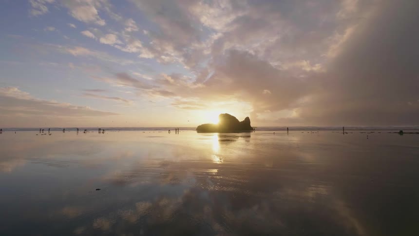 The shore has a large rock and the sun is setting behind it