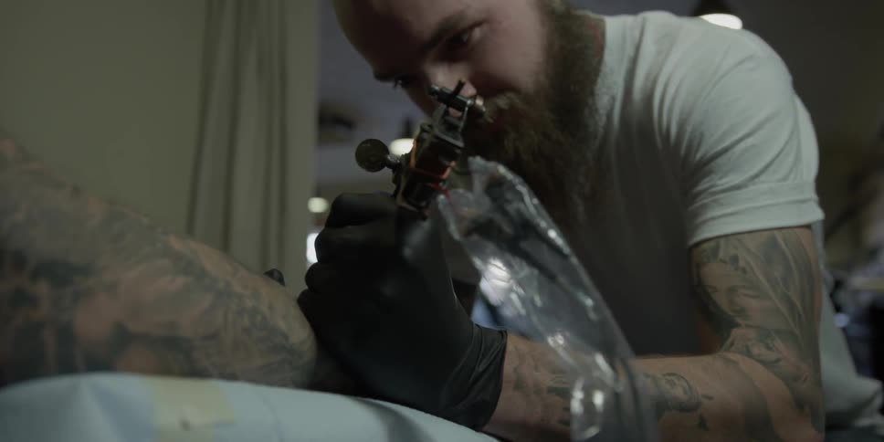A tattoo artist is working on a client in a shop during the day