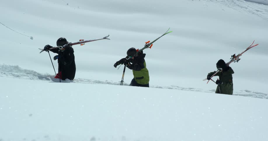 Skiers hike up snowy mountain with skis on shoulders