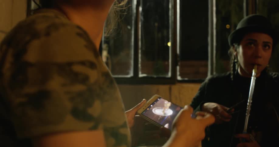 A woman uses her phone as another smokes from a pipe