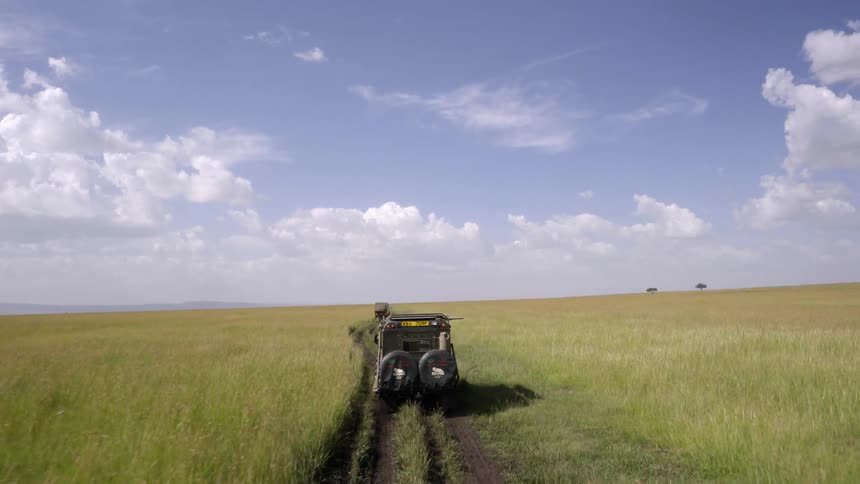 Two jeeps travel on a dirt path across an african savanna during the day