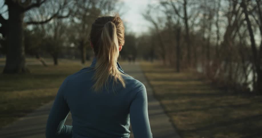 Blonde woman with pony tail and a blue jacket jogs along walking trail in a public park
