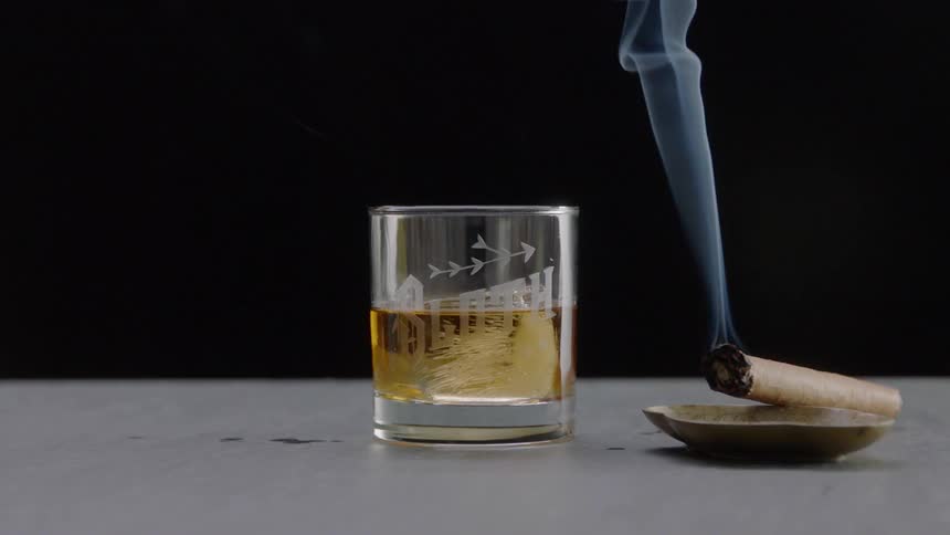 Glass of whiskey rests next to a burning cigar