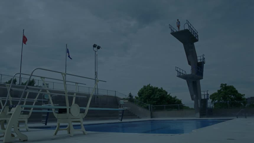 Someone walks to the ledge and jumps off the highest platform diving board into a pool below