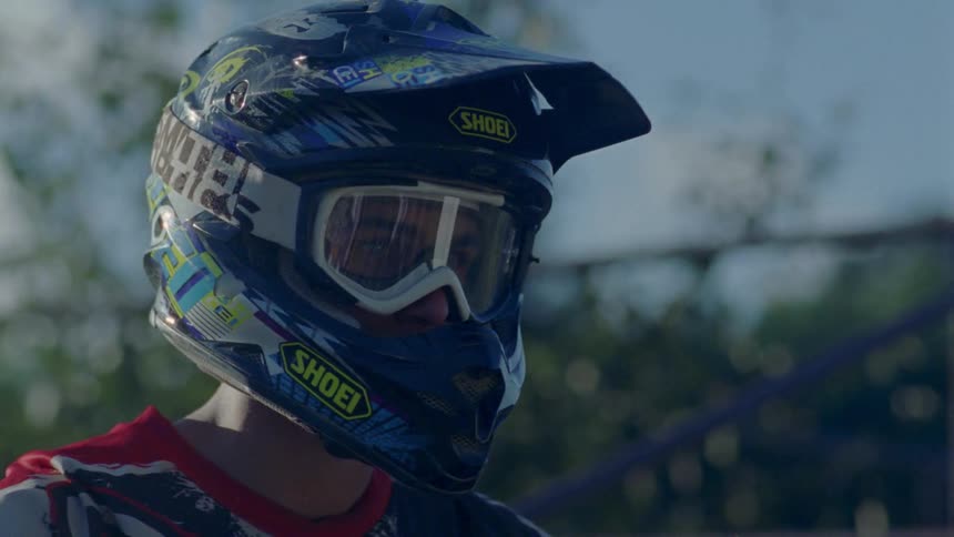 A man with a motorcycle helmet and goggles on is standing around looking at another rider