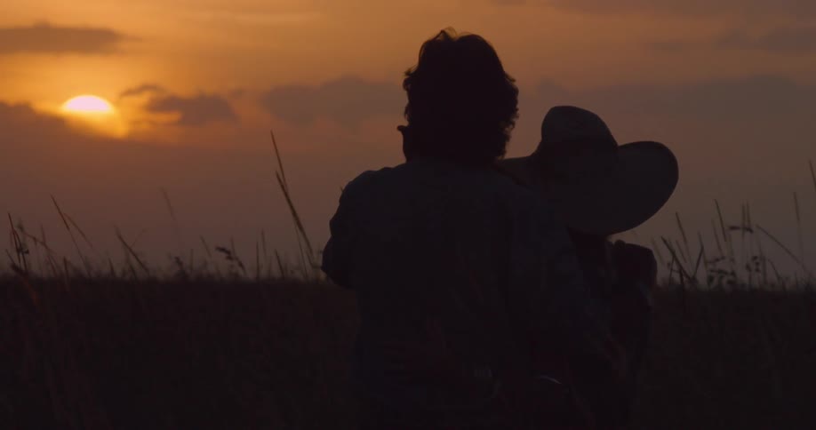 A couple hold onto each other as they watch the sun setting over a grassy plain