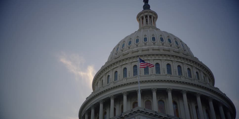 An american flag billows in front of a capitol building