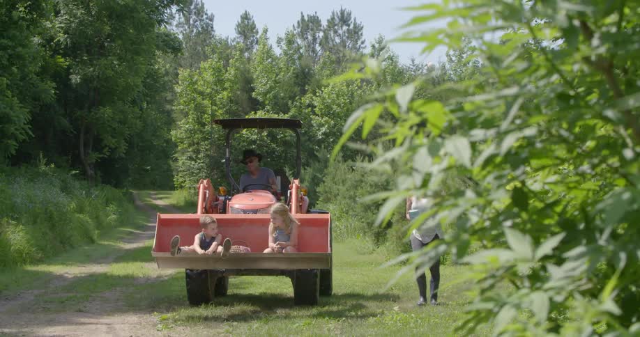Two kids are sitting in the shovel of a tractor while someone drives it and someone else walks alongside