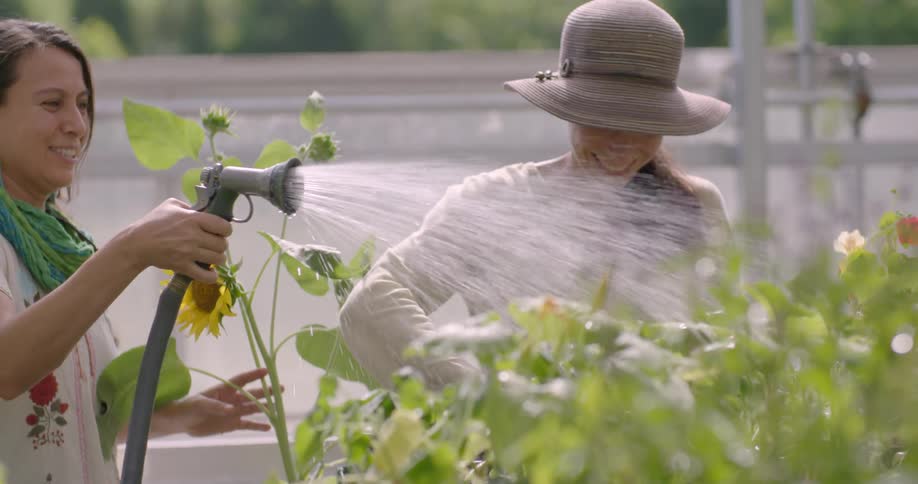 Two women laugh and talk as they water plants in a garden