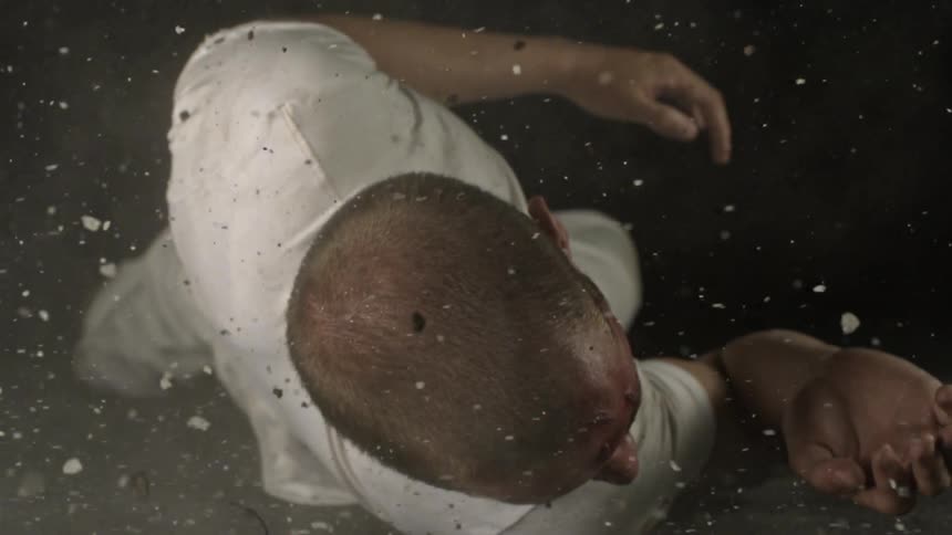 A man is rolling on the ground as bits of rock and debris hit him