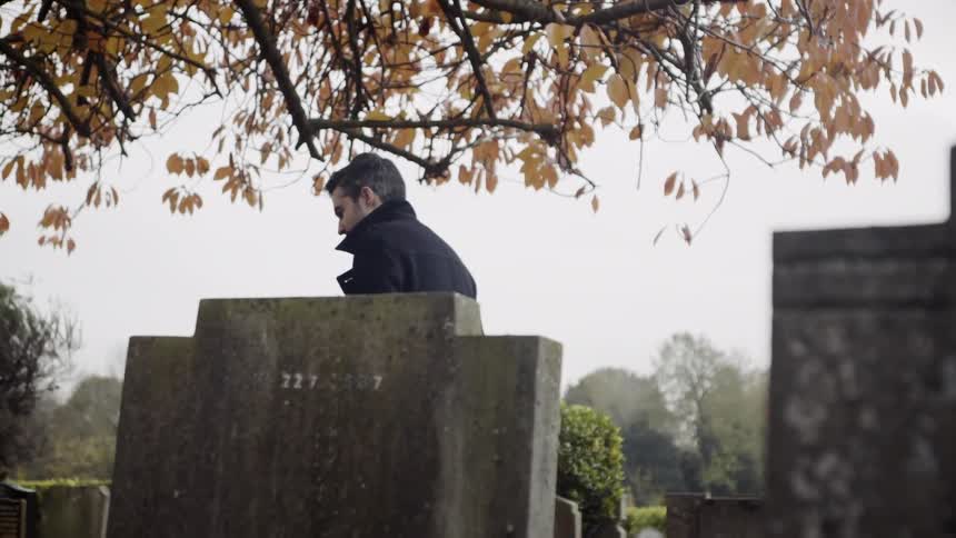 A man in a pea coat is looking at the tombstone of someone and then walks away