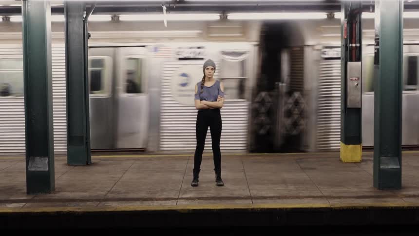 A train passes behind a woman standing waiting at a subway station