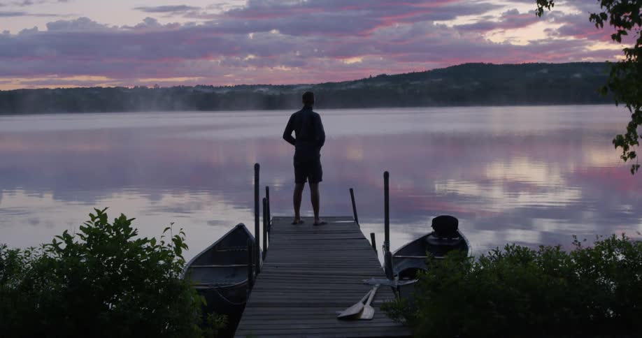 A man stands on the end of a dock watching the sunset behind the mountains