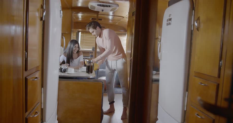 A man is pouring orange juice for a woman sitting at a table in a trailer