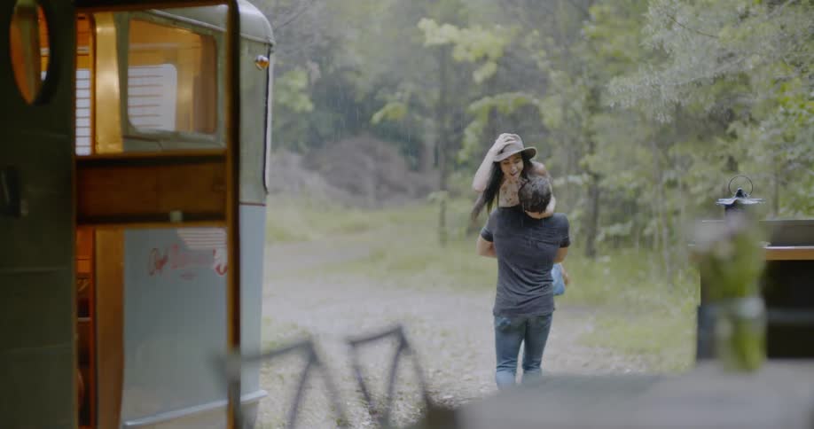 A man is lifting and carrying a woman outside a camper in a natural setting