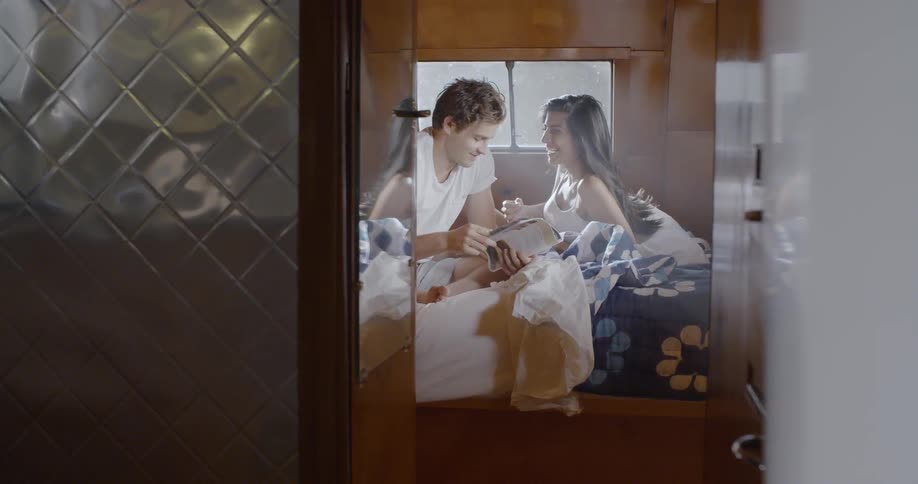 A man and a woman rest on a bed in a trailer reading and laughing