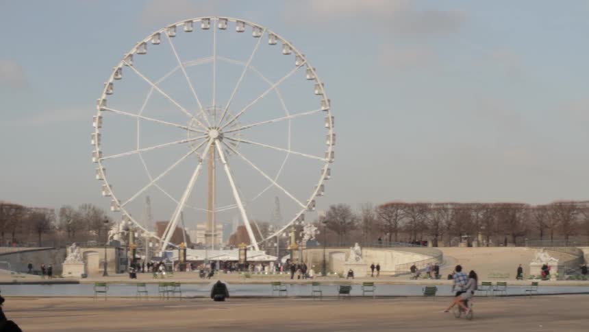 A couple ride a bike in a park mall with a giant ferris wheel in the background