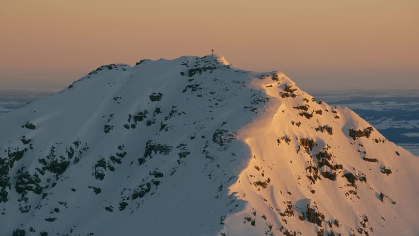 A cross stands atop a mountain peak covered in snow at dusk