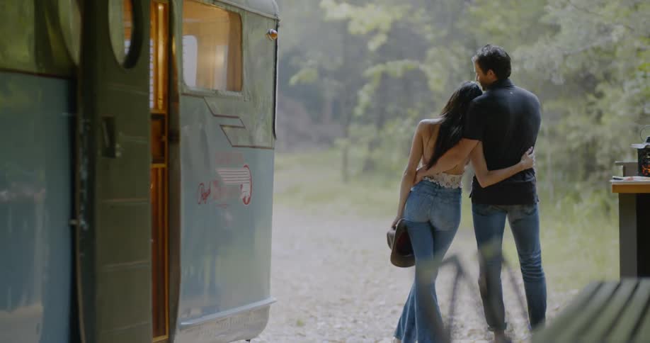 A woman removes her hat as she stands with a man outside a camper trailer