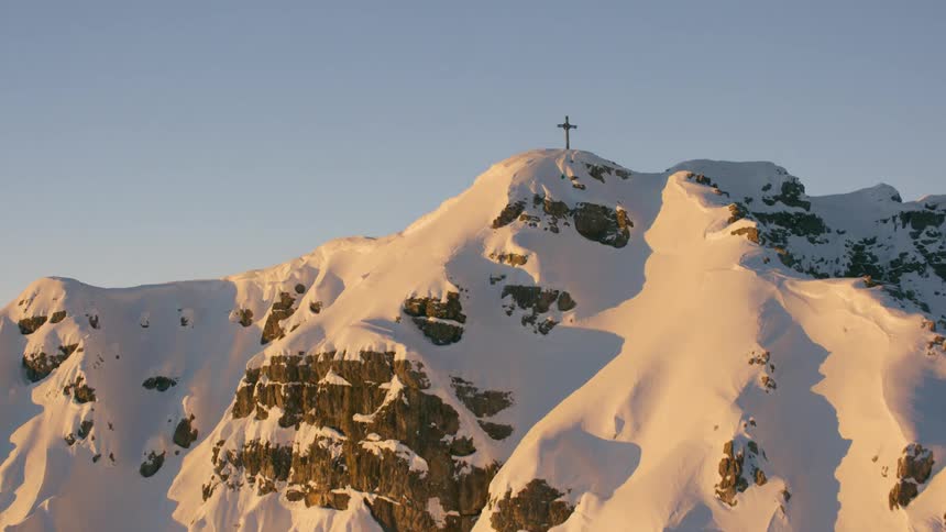 A cross stands atop a mountain peak covered in snow at dawn