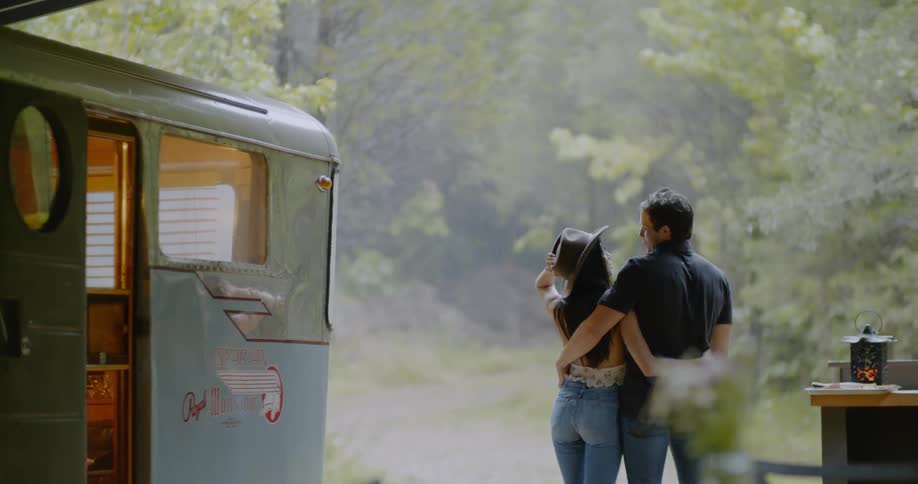 A man and woman stand next to a camper in the woods