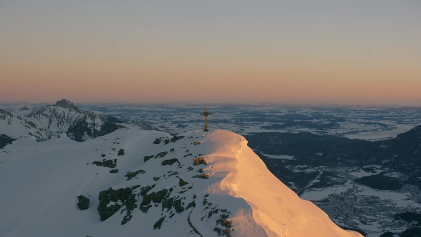 A cross stands atop a mountain peak covered in snow at dusk