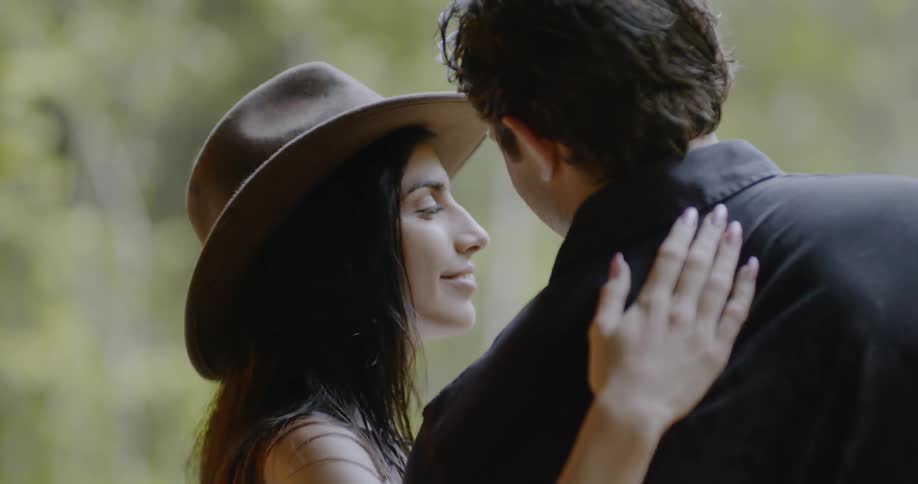 A woman caresses the back of a man's head as they stand outside during the day