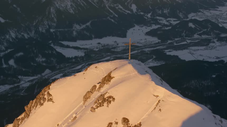 A cross stands atop a mountain peak covered in snow at dusk