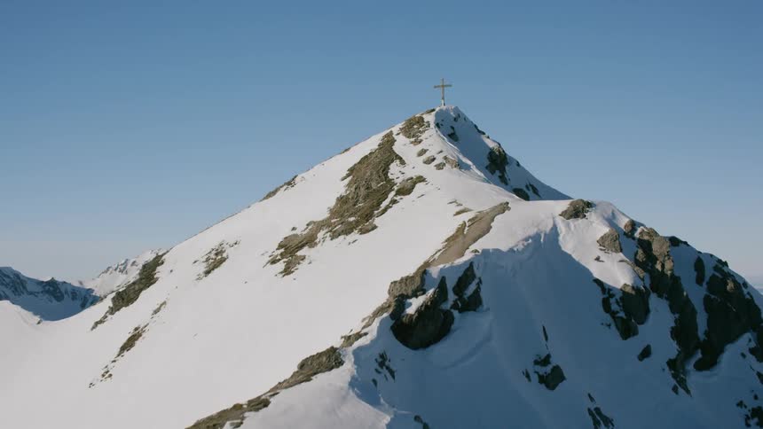 A cross stands atop a mountain peak covered in snow on a sunny day