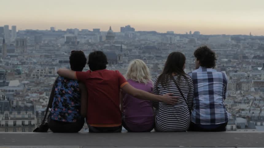 A group of men and women chat and laugh while looking out over a cityscape as they sit together with their legs dangling off the top of building