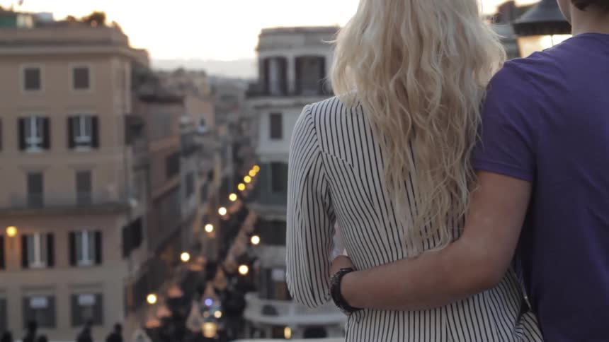 A white man and white woman stand on a rooftop in a city neighborhood and watch whats going on down on the street
