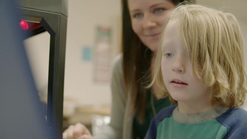 A woman is watching a child use a 3d printer in a classroom