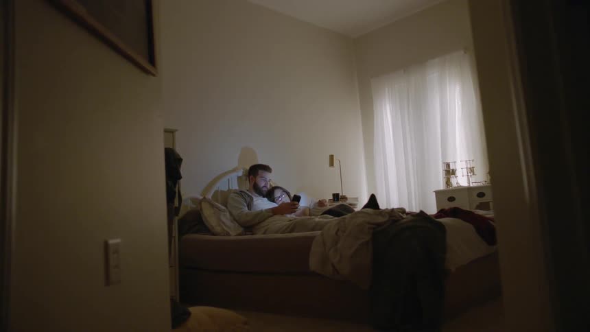 A man and woman are laying in bed together looking at a cellphone screen