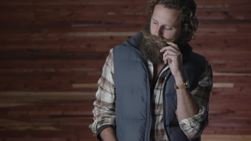 Man with a grey vest and long hair touches his long beard