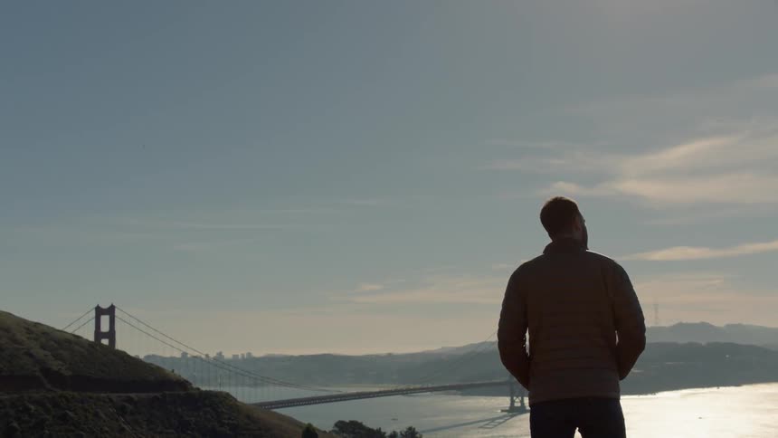 Man standing on a hill overlooking a large river and bridge