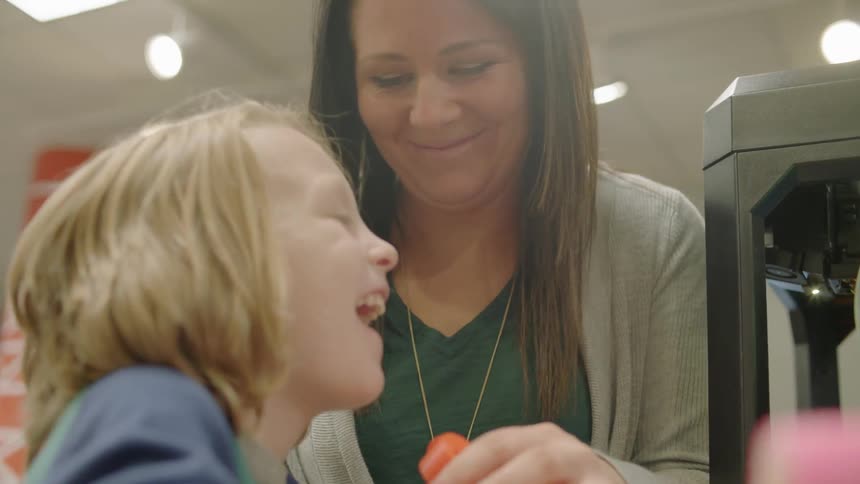 Child smiles and holds and orange plastic object while a woman stands over him smiling