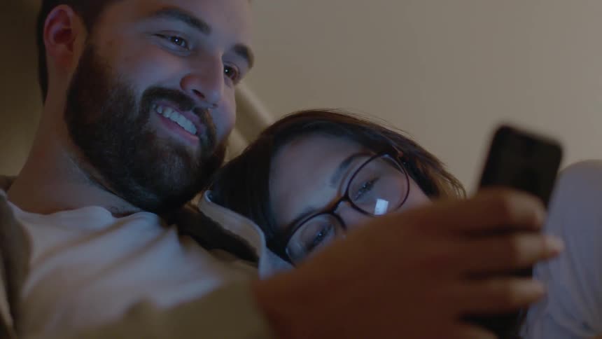 A man and woman cuddle in bed while looking at a phone