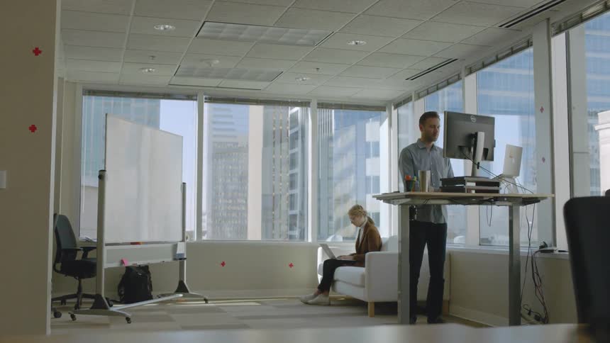 A man is at a standing desk as a woman works on the couch