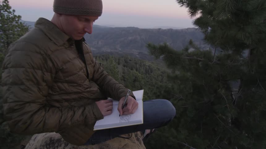 A white sits on a mountain and writes in a notebook