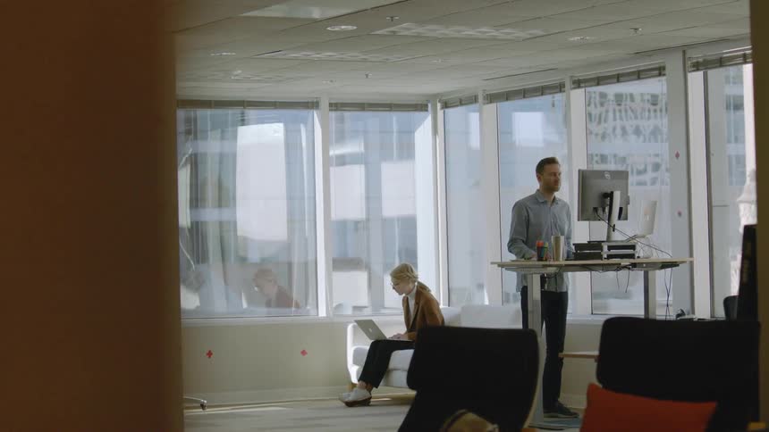 A man is standing at computer while another woman is sitting using a laptop behind him