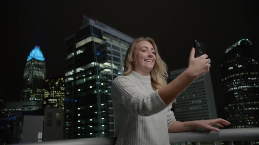 A woman is standing on a balcony taking a selfie with the city behind her