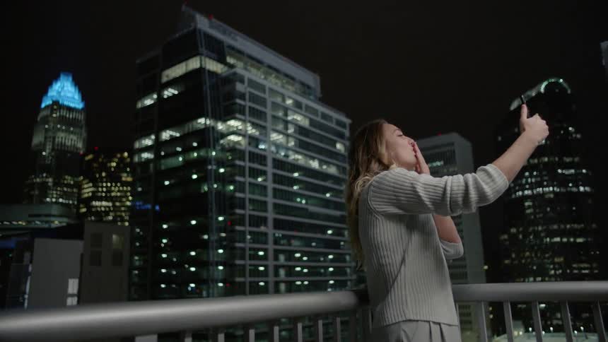 A woman is smiling as she takes pictures of herself and a city skyline at night