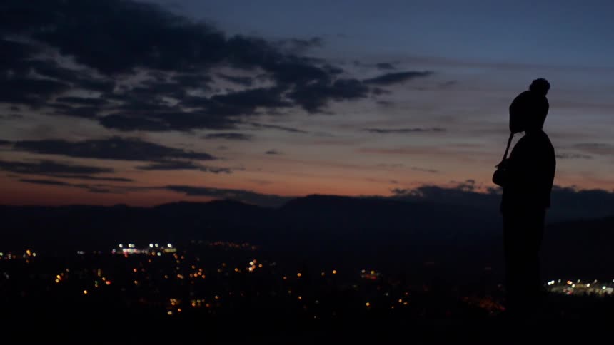 Person standing on hill above city lights at sunset