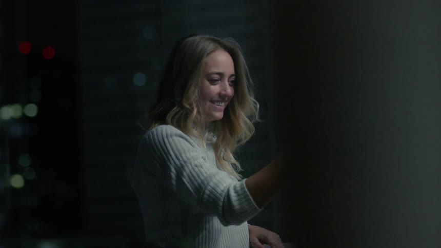 Woman on balcony in city at night smiling and laughing during video call