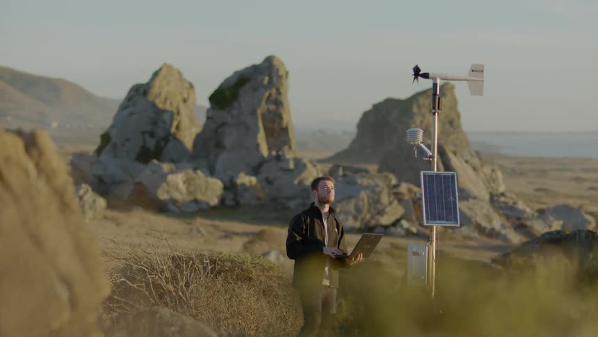 Man stands outside holding laptop and looking at a small metal machine