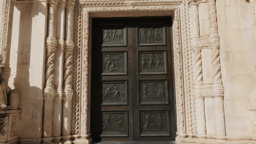 A ornate door with pictures on it is connected to an old white stone building