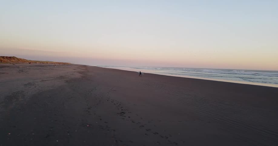 Person carries a surfboard across a beach at sunset